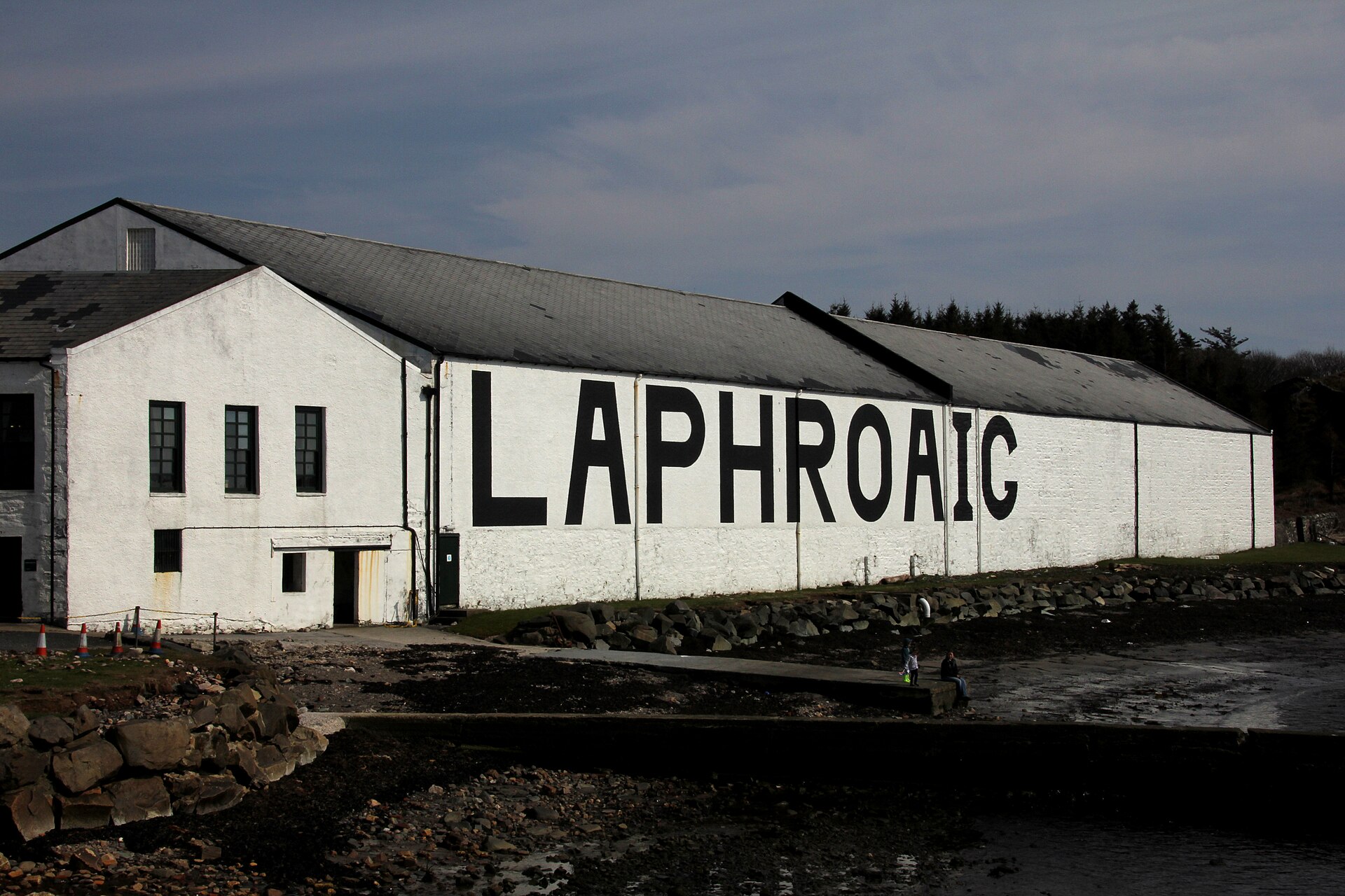 The whitewashed seafront warehouses of Laphroaig distillery on Islay under a dark sky, seen across the rocky shore